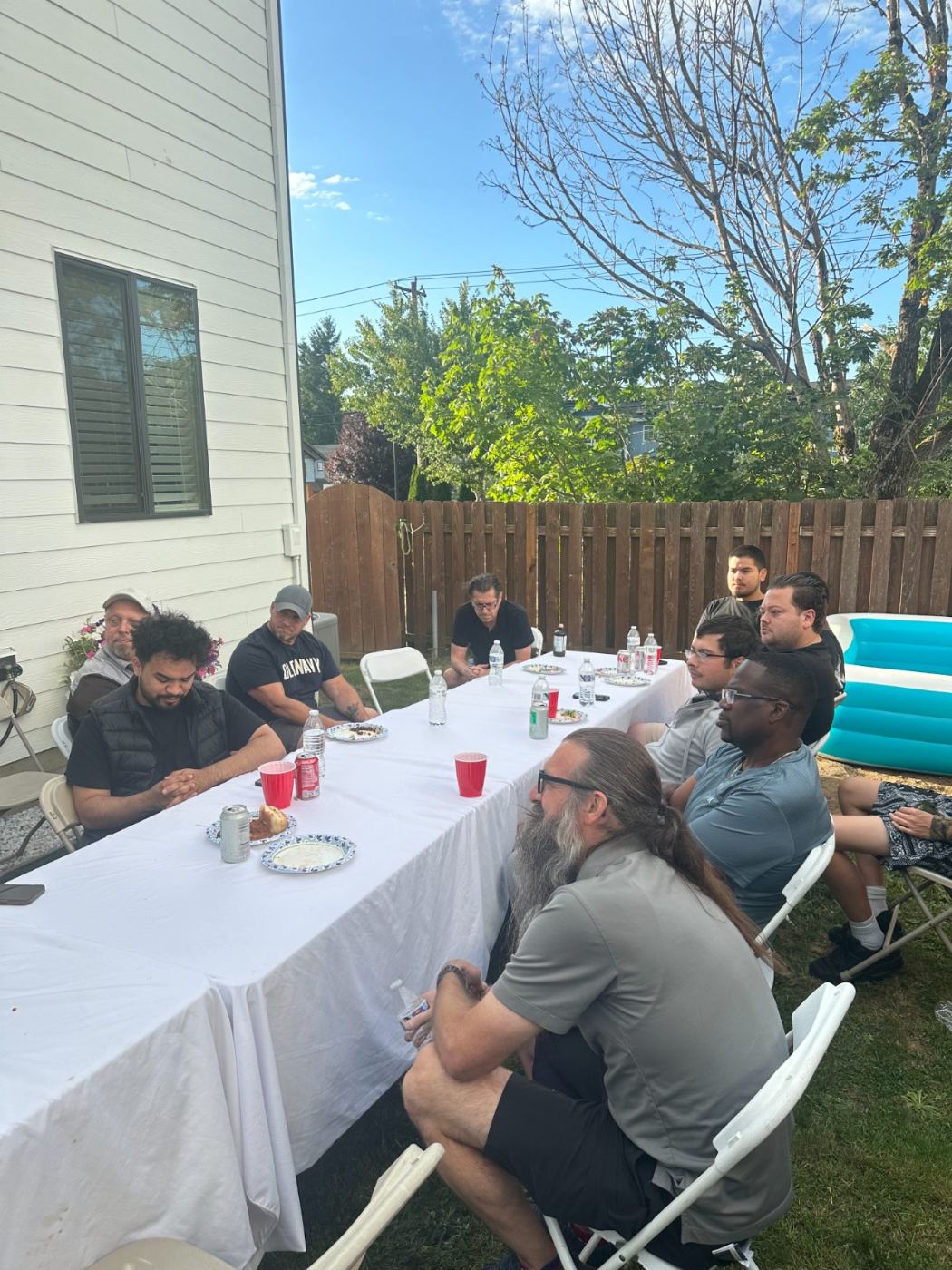 Men standing in prayer at a backyard table, one with a hand raised