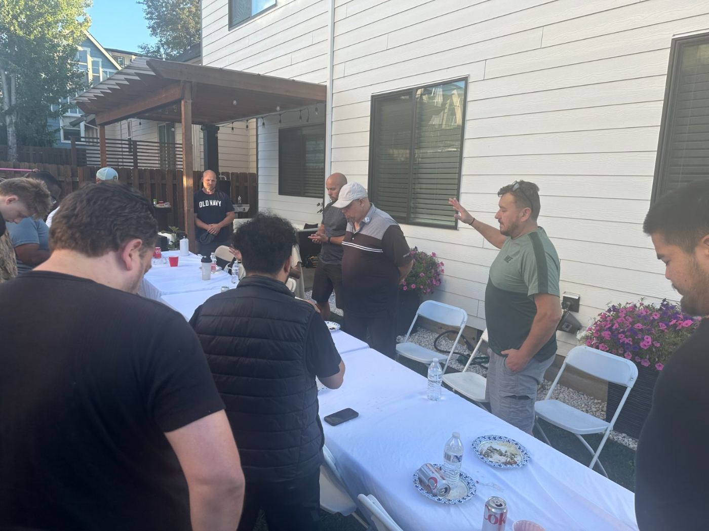 Men seated along a long table in a backyard, listening together