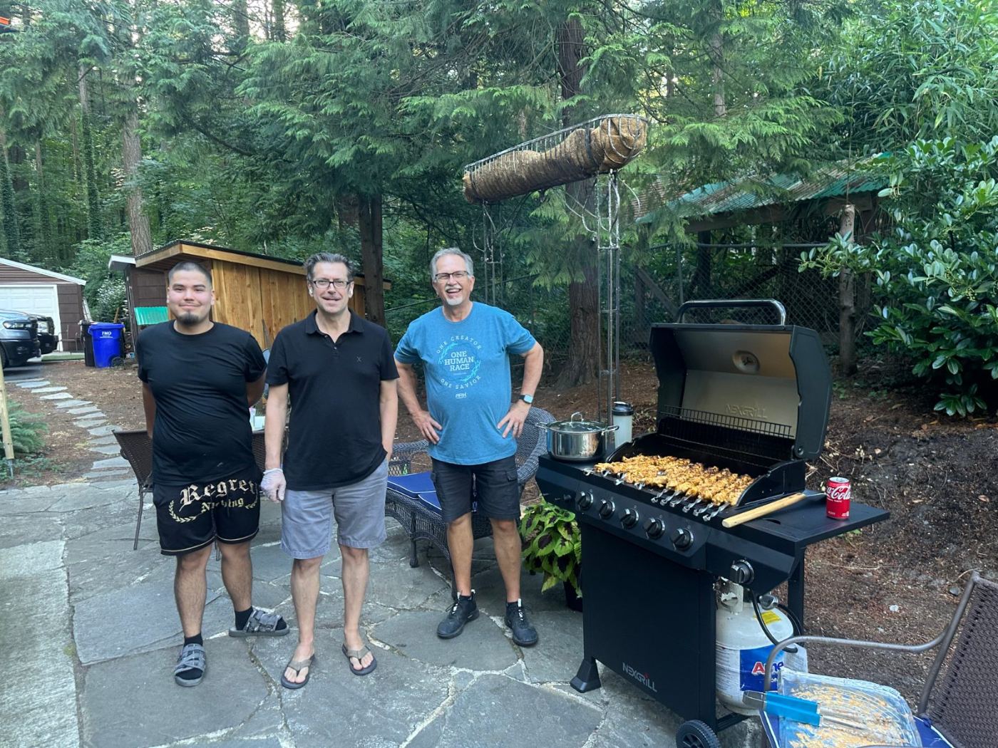 Three men smiling beside a grill lined with chicken skewers