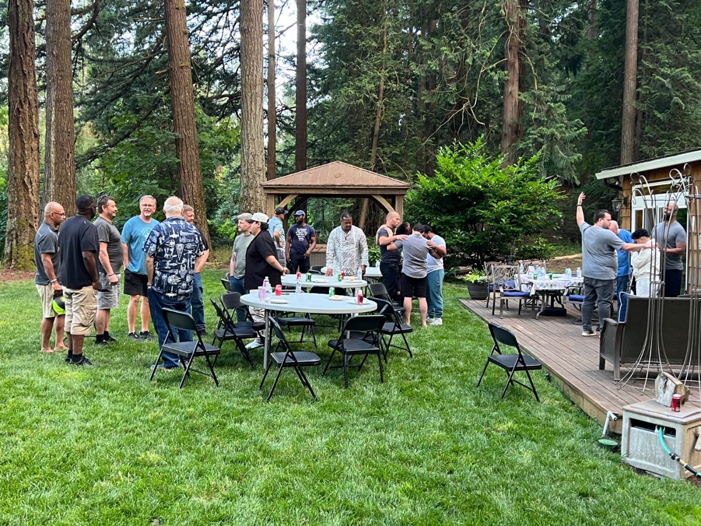 A large group of men fellowshipping around tables in a forest backyard