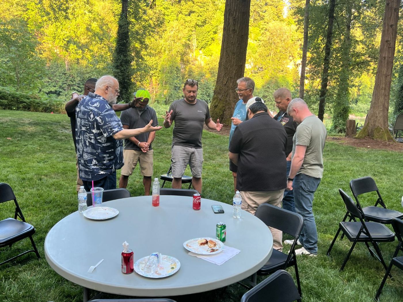 Men standing in prayer around a table as evening light falls through the trees